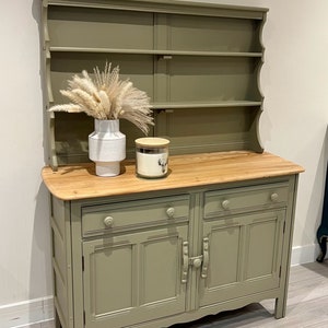 May include: A green painted wooden hutch with a top shelf and two lower cabinets. The hutch has a wood top and a white vase with dried pampas grass.