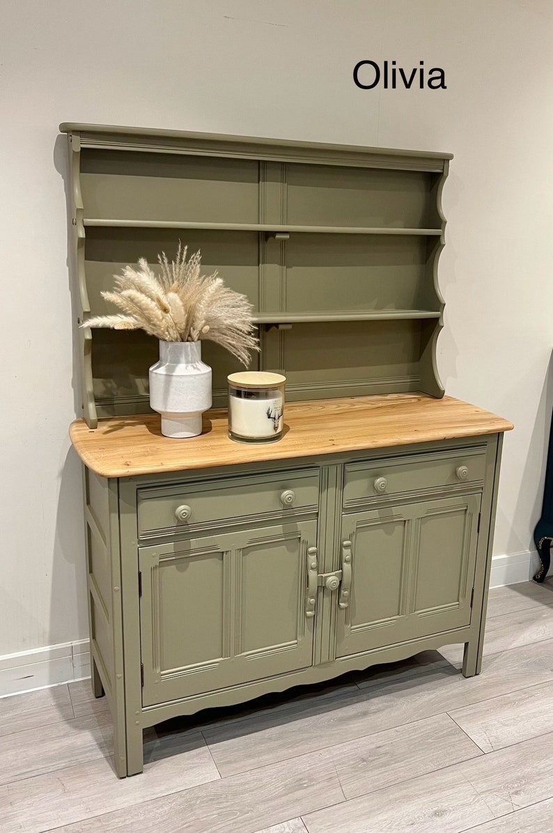 May include: A green painted wooden hutch with a top shelf and two lower cabinets. The hutch has a wood top and a white vase with dried pampas grass.