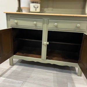 May include: A sage green hutch with two drawers and two cabinet doors. The hutch has a top shelf and a wooden countertop. The cabinet doors are open, revealing the interior shelves.