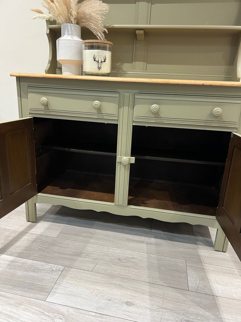 May include: A sage green hutch with two drawers and two cabinet doors. The hutch has a top shelf and a wooden countertop. The cabinet doors are open, revealing the interior shelves.