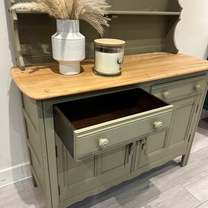 May include: A sage green painted hutch with a wooden top and a single drawer. The hutch has two doors with decorative details and a top shelf. The drawer is open and the interior is visible.