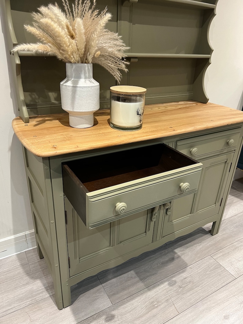 May include: A sage green painted hutch with a wooden top and a single drawer. The hutch has two doors with decorative details and a top shelf. The drawer is open and the interior is visible.