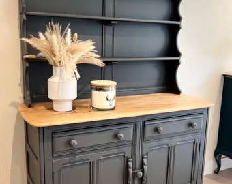 May include: A dark grey painted dresser with a wooden top and two doors. The dresser has a top shelf with two additional shelves below. The dresser has a white vase with dried pampas grass and a glass jar with a candle inside.