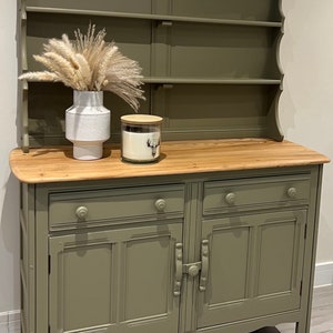 May include: A green painted wooden hutch with a top shelf and two doors. The hutch has a wooden top and two drawers. The hutch is sitting on a wooden floor.