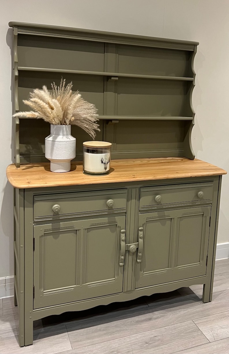 May include: A green painted wooden hutch with a top shelf and two doors. The hutch has a wooden top and two drawers. The hutch is sitting on a wooden floor.