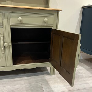 May include: A light green painted wooden hutch with a wood top and a single drawer. The hutch has a cabinet with a door that is open, revealing a shelf inside. The cabinet has a wood interior.