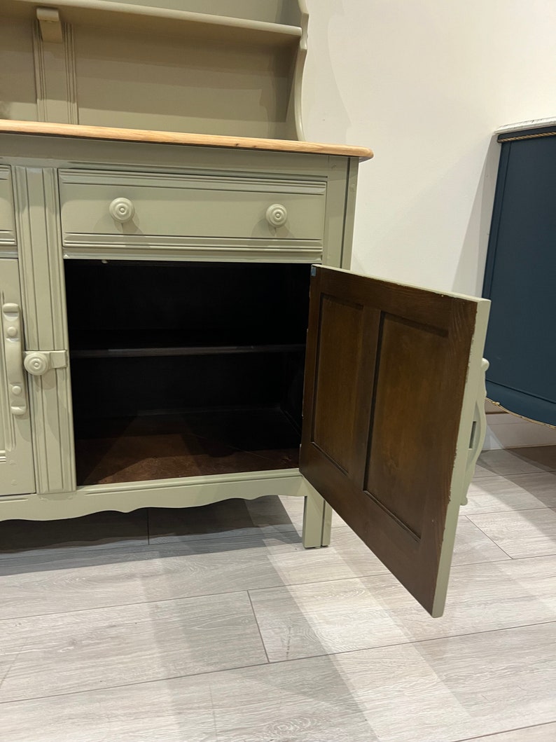 May include: A light green painted wooden hutch with a wood top and a single drawer. The hutch has a cabinet with a door that is open, revealing a shelf inside. The cabinet has a wood interior.