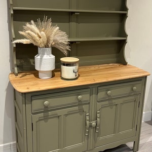 May include: A green painted hutch with a wooden top and two doors. The hutch has a shelf on the top and a drawer below the doors. There is a white vase with dried pampas grass and a candle on the top.