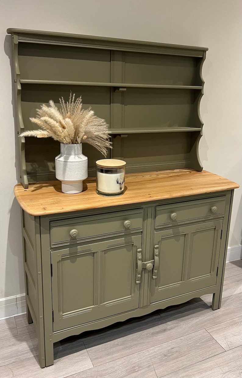 May include: A green painted hutch with a wooden top and two doors. The hutch has a shelf on the top and a drawer below the doors. There is a white vase with dried pampas grass and a candle on the top.