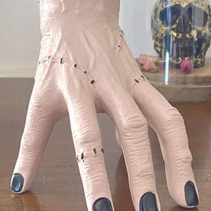 May include: A pale pink ceramic hand with black fingernails and stitched details. The fingers are slightly curved, and the wrist is open. The hand is a decorative item, possibly a vase or holder, with a spooky or gothic aesthetic.