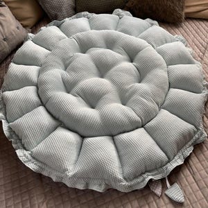 Pu&ograve; includere: Un cuscino da pavimento rotondo di colore azzurro con una texture a nido d'ape e un bordo arricciato. Il cuscino &egrave; riempito con pi&ugrave; strati di imbottitura, creando una superficie comoda e di supporto.
