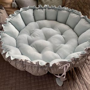 Pu&ograve; includere: Un letto rotondo per animali domestici di colore blu chiaro e grigio con un bordo arricciato e un fiocco bianco. Il letto &egrave; riempito con un'imbottitura morbida e presenta un design trapuntato.