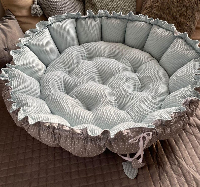 Pu&ograve; includere: Un letto rotondo per animali domestici di colore blu chiaro e grigio con un bordo arricciato e un fiocco bianco. Il letto &egrave; riempito con un'imbottitura morbida e presenta un design trapuntato.