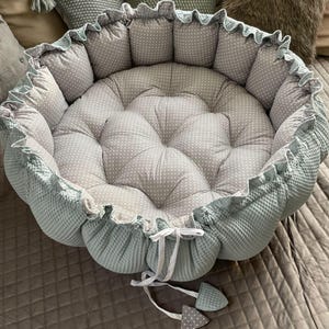 Pu&ograve; includere: Un letto rotondo per animali domestici di colore verde chiaro e grigio con un volant bianco attorno al bordo. Il letto ha un design trapuntato con pois bianchi sul tessuto grigio.