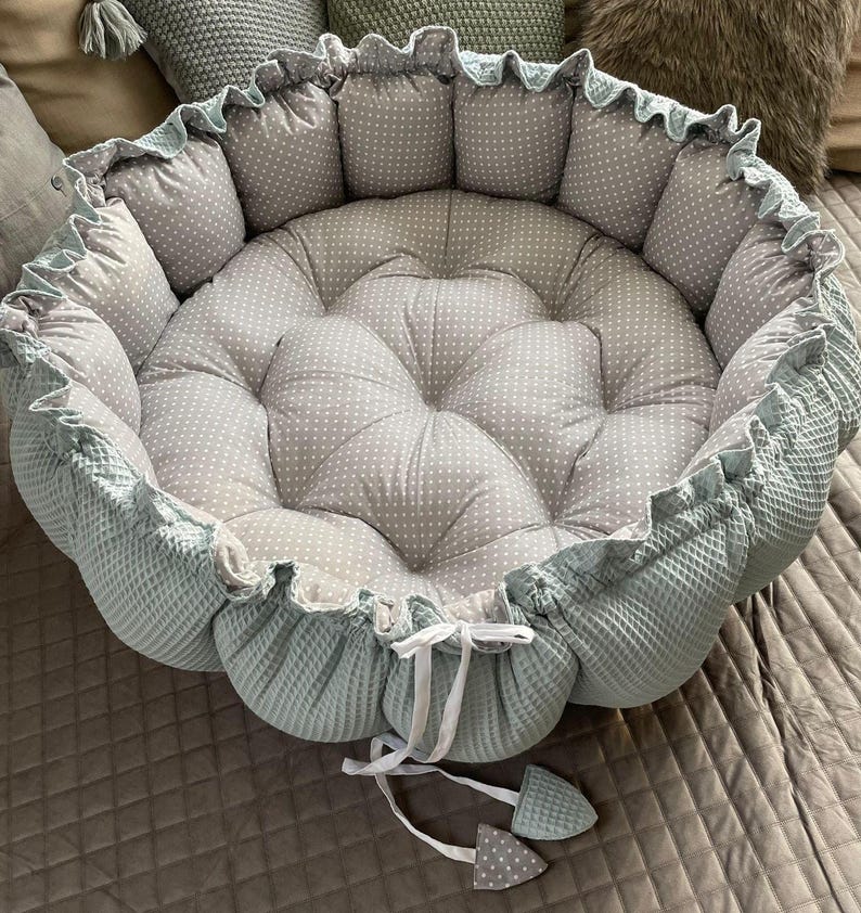 Pu&ograve; includere: Un letto rotondo per animali domestici di colore verde chiaro e grigio con un volant bianco attorno al bordo. Il letto ha un design trapuntato con pois bianchi sul tessuto grigio.