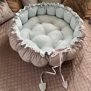 Pu&ograve; includere: Un nido rotondo per neonati di colore blu chiaro e grigio con un nastro bianco e due nappine bianche. Il nido &egrave; realizzato in tessuto morbido e presenta un design trapuntato.