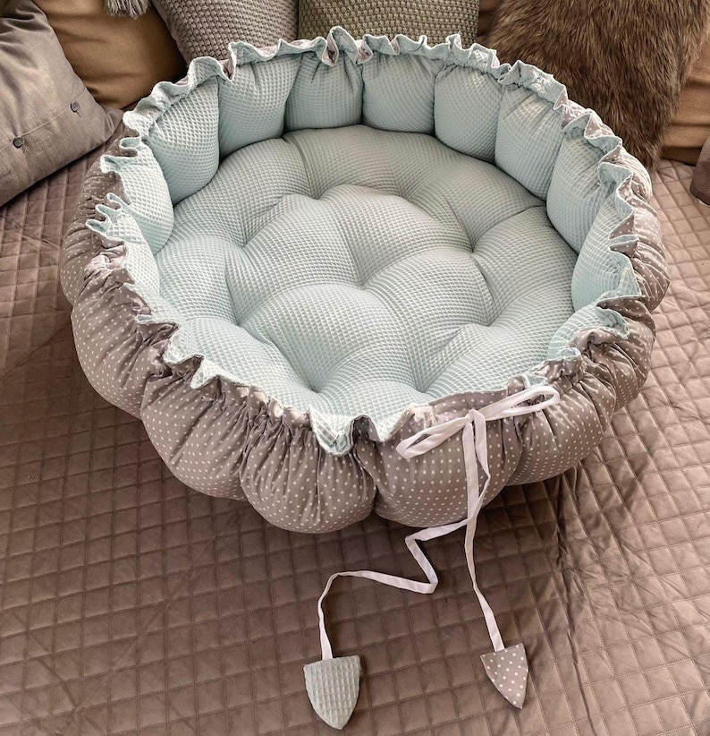 Pu&ograve; includere: Un nido rotondo per neonati di colore blu chiaro e grigio con un nastro bianco e due nappine bianche. Il nido &egrave; realizzato in tessuto morbido e presenta un design trapuntato.