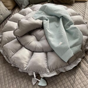Pu&ograve; includere: Un cuscino rotondo grigio e bianco a pois con una coperta in tessuto a nido d'ape blu chiaro sopra. Il cuscino ha un bordo arricciato ed &egrave; riempito con un'imbottitura morbida.