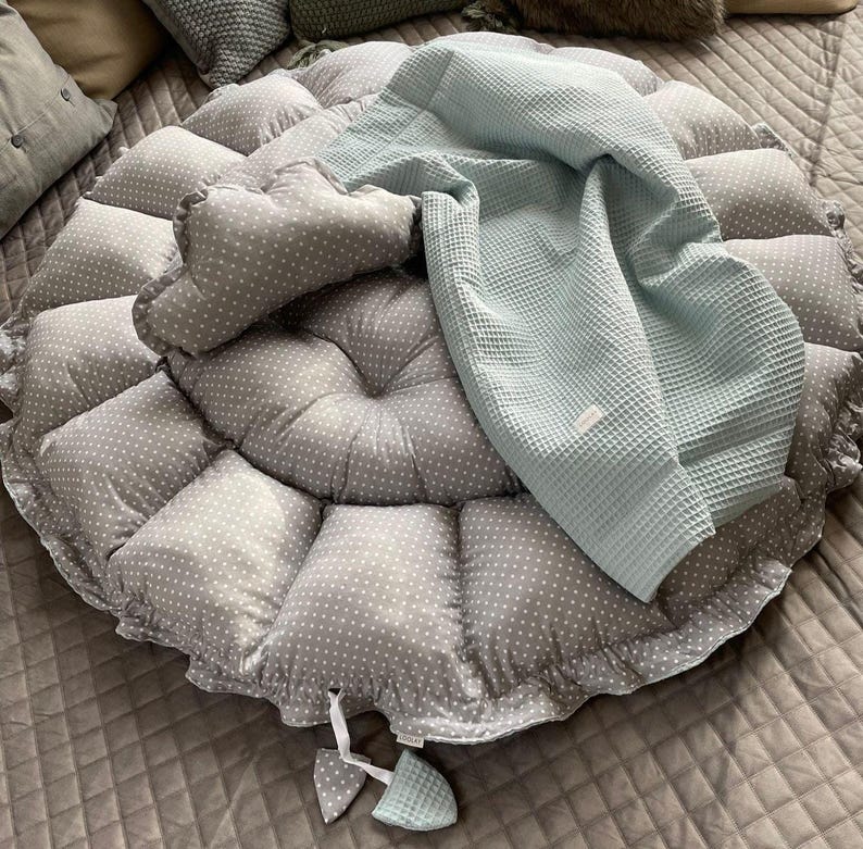 Pu&ograve; includere: Un cuscino rotondo grigio e bianco a pois con una coperta in tessuto a nido d'ape blu chiaro sopra. Il cuscino ha un bordo arricciato ed &egrave; riempito con un'imbottitura morbida.