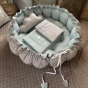 Op de afbeelding: Een grijs en wit gestippeld baby nest met een lichtblauw gestikt dekentje en kussen. Het nest heeft een witte strik.