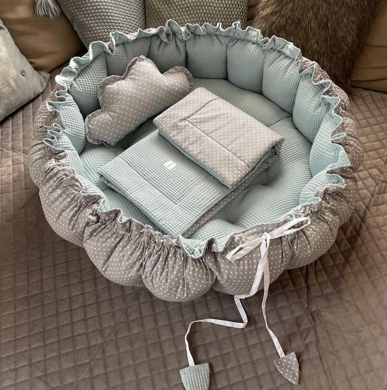 Pu&ograve; includere: Un nido per neonati grigio e bianco a pois con una coperta trapuntata blu chiaro e un cuscino. Il nido ha un nastro bianco.