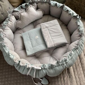 Pu&ograve; includere: Un nido per neonati di colore blu chiaro e grigio con un motivo a pois bianchi. Il nido ha un cuscino rimovibile e una coperta abbinata.