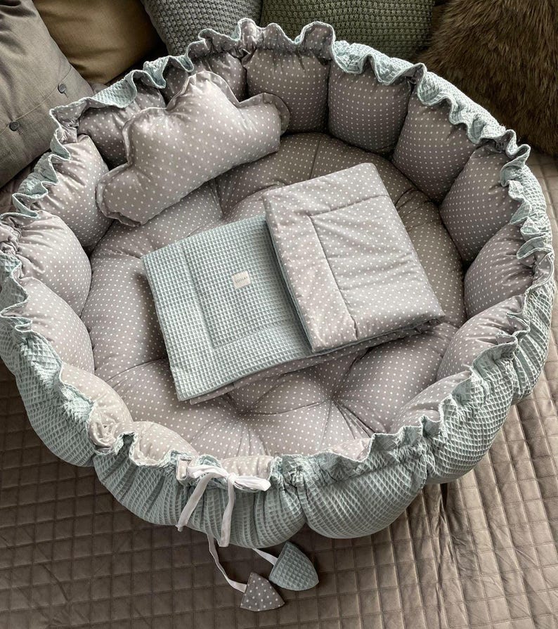 Pu&ograve; includere: Un nido per neonati di colore blu chiaro e grigio con un motivo a pois bianchi. Il nido ha un cuscino rimovibile e una coperta abbinata.
