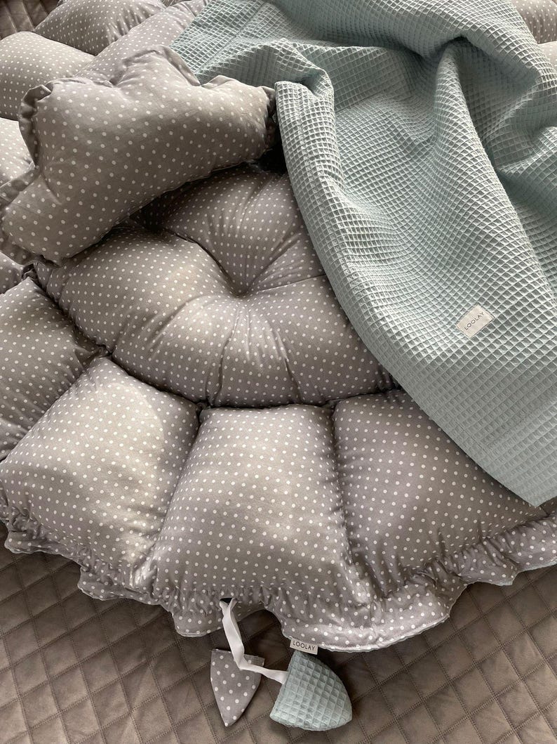 Pu&ograve; includere: Un nido per neonati grigio e bianco a pois con una coperta in tessuto a nido d'ape blu chiaro e un giocattolo blu chiaro abbinato. Il nido ha un bordo arricciato e la coperta ha la scritta "LOOLAY" stampata.
