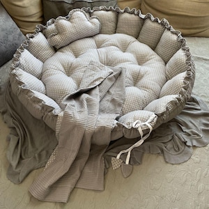 Op de afbeelding: Een rond grijs en wit gestippeld kussen met een grijs wafel geweven deken en een grijs kussen. Het kussen heeft een gerimpelde rand en is gevuld met knopen.