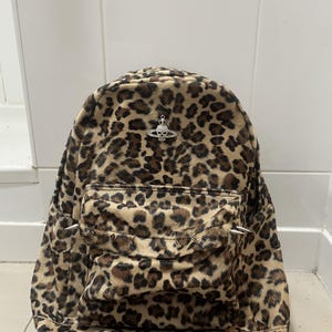 Könnte beinhalten: Ein Rucksack mit Leopardenmuster und einer weichen, strukturierten Oberfläche. Die Vordertasche ist mit einer Reihe silberfarbener Metallspitzen verziert. Über der Tasche befindet sich ein silberfarbenes Metallemblem in Planetenform mit Totenkopf.