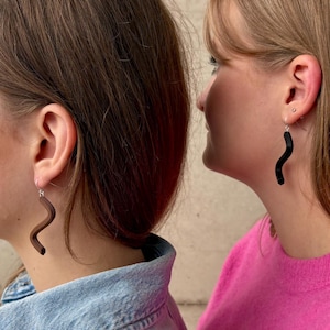Könnte beinhalten: Zwei Frauen mit wellenförmigen, abstrakten Ohrringen. Eine Frau trägt eine Jeansjacke, die andere einen rosa Pullover. Die Ohrringe sind dunkel und haben eine geschwungene, längliche Form. Der Hintergrund ist eine neutrale, strukturierte Wand.