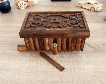 May include: A wooden box with a carved floral design on the lid. The box has a secret opening mechanism and a small metal ring on the side.