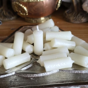 May include: A collection of small, cylindrical off-white wax candles with visible wicks, piled together on a metallic, triangular-shaped tray. The candles have a smooth, solid texture and are arranged in a cluster, showing multiple individual units.