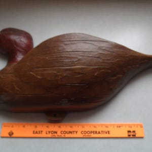 May include: A wooden duck decoy with a brown body and a red head. The decoy has carved details and a smooth finish. An orange ruler with text is at the bottom of the image.