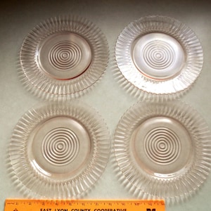 Puede incluir: Cuatro platos de vidrio transparente con un diseño en espiral en el centro y un borde acanalado. Los platos están dispuestos en forma cuadrada. Los platos son de color rosa claro.