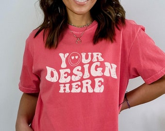 May include: A woman wearing a coral colored t-shirt with the text "YOUR DESIGN HERE" in white with a smiley face and hearts. The shirt is a relaxed fit and has a distressed look.
