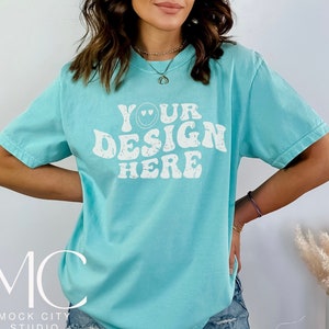 Puede incluir: Una camiseta azul turquesa con un gráfico blanco que dice "Your Design Here" en una fuente retro con una carita sonriente.