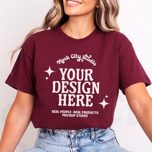 Può includere: Una maglietta color bordeaux con maniche corte che presenta il testo "Mock City Studio", "Your Design Here", "Real People - Real Products" e "Mockup Studio".