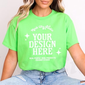May include: A woman wearing a bright green t-shirt with the text "Mock City Studio" and "Your Design Here" in white. The shirt also has the text "Real People. Real Products. Mockup Studio" in white. She is wearing blue jeans.