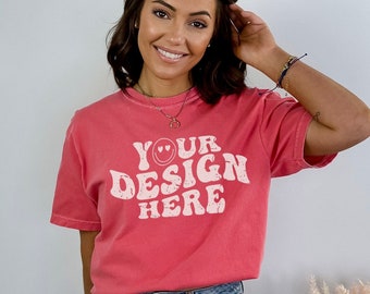 May include: A woman wearing a coral colored t-shirt with the text "YOUR DESIGN HERE" in white. The text is in a retro font with a smiley face and hearts. She is wearing blue denim shorts.