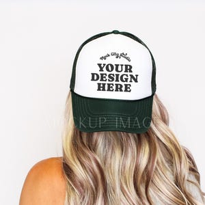 Könnte beinhalten: Dunkelgrüne und weiße Trucker-Mütze mit dem Text "YOUR DESIGN HERE" in Schwarz. Die Mütze wird von einer Person mit blonden und braunen Haaren getragen. Die Mütze hat einen Mesh-Rücken.
