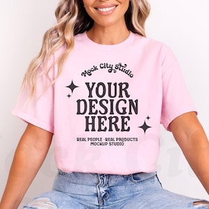 May include: A pink t-shirt with the text "Mock City Studio" and "Your Design Here" in black. The text is in a retro font style with stars on either side. The text below reads "Real People - Real Products Mockup Studio".