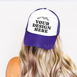 May include: A purple and white trucker hat with the text "YOUR DESIGN HERE" in black. The hat has a mesh back and a curved brim. The hat is worn by a person with long, wavy blonde hair.
