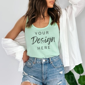 Puede incluir: Una mujer lleva una camiseta sin mangas de color verde claro con el texto "YOUR Design HERE" impreso en la parte delantera. También lleva pantalones cortos de mezclilla y una camisa blanca abotonada.