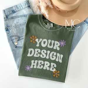 Puede incluir: Una camiseta verde con un gráfico blanco que dice "YOUR DESIGN HERE" y tiene flores naranjas y moradas alrededor del texto.