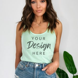Könnte beinhalten: Hellgrünes Tanktop mit dem Text "YOUR Design HERE" in schwarzer Schrift. Das Model trägt Jeansshorts und eine goldene Halskette. Der Hintergrund ist eine schlichte weiße Wand mit einer grünen Pflanze.