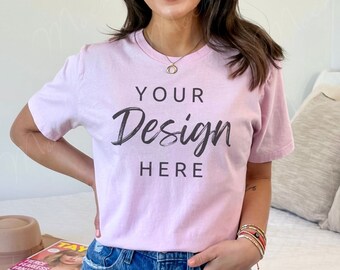 Puede incluir: Una camiseta rosa claro con el texto "YOUR Design HERE" en escritura negra. La modelo lleva vaqueros azules. Un sombrero marrón y una revista están en la cama. La cama tiene un edredón blanco y almohadas beige.