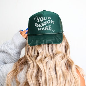 Könnte beinhalten: Dunkelgrüne Trucker-Mütze mit dem Text "YOUR DESIGN HERE" in weißer Schrift. Die Mütze hat eine Netzrückseite und einen gebogenen Schirm. Die Mütze wird von einer Person mit langen, welligen blonden Haaren getragen.