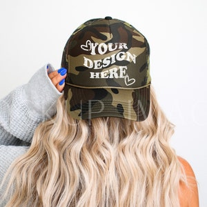 May include: A camouflage trucker hat with white text that says "YOUR DESIGN HERE" and two small hearts on either side of the text.