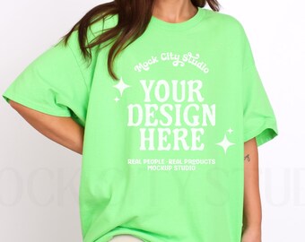 Puede incluir: Una camiseta de gran tamaño de color verde brillante con el texto "Mock City Studio" y "YOUR DESIGN HERE" en blanco. El texto está rodeado de pequeñas estrellas blancas. El texto debajo del diseño dice "REAL PEOPLE-REAL PRODUCTS MOCKUP STUDIO".
